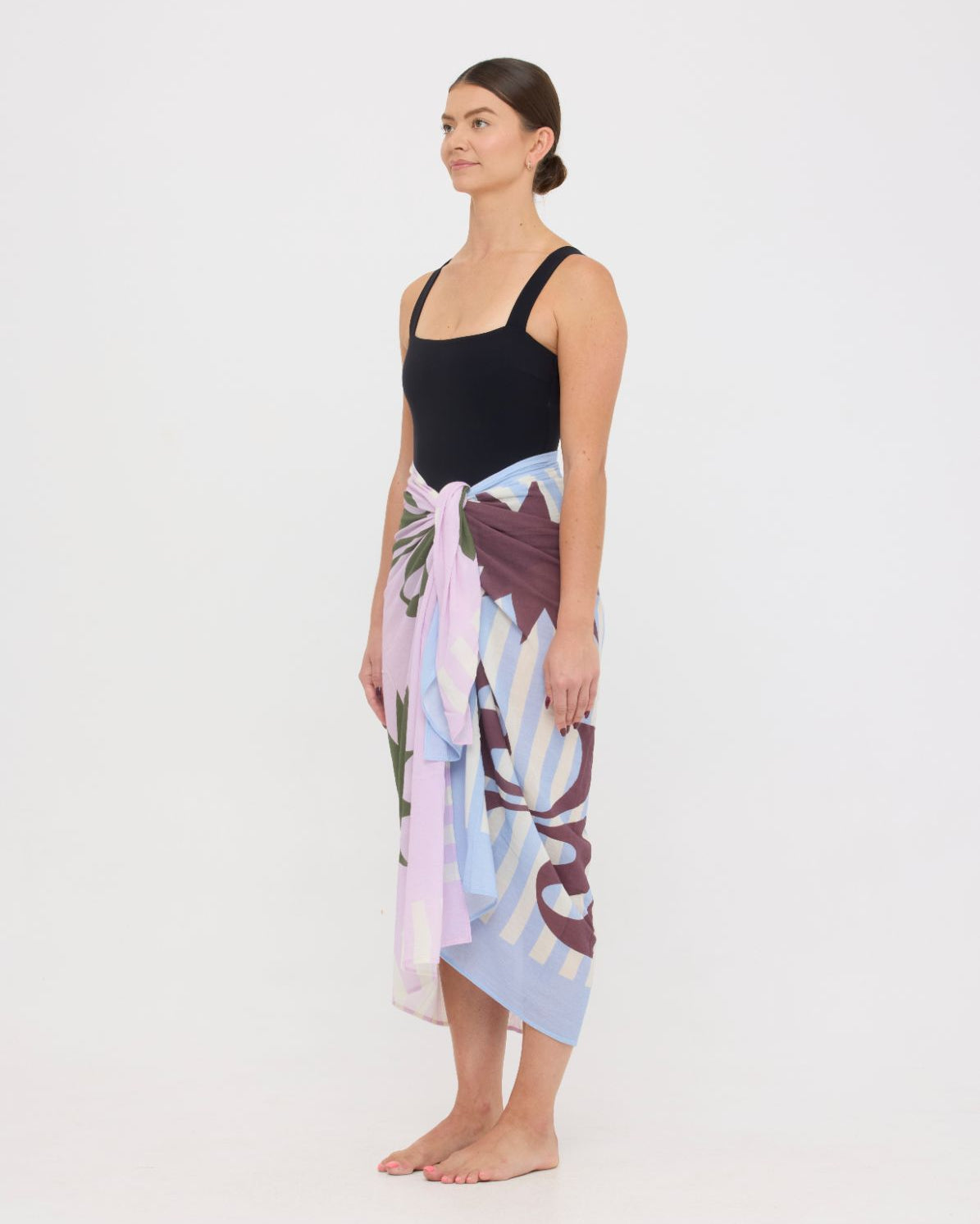 Woman wearing the black HUZZAH Scout sculpting one-piece with a lightweight multi coloured sarong wrapped at the hip, creating a sophisticated swimwear look.