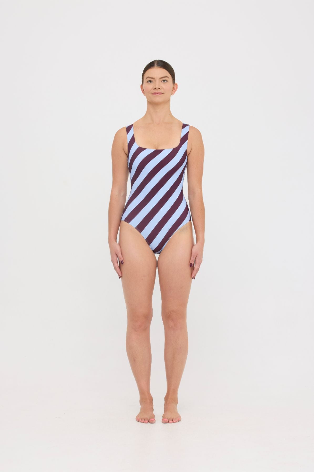 Model wearing the dusty blue and plum rising stripe HUZZAH Cat one-piece swimsuit with sculpting fit and clean lines, photographed against a plain background