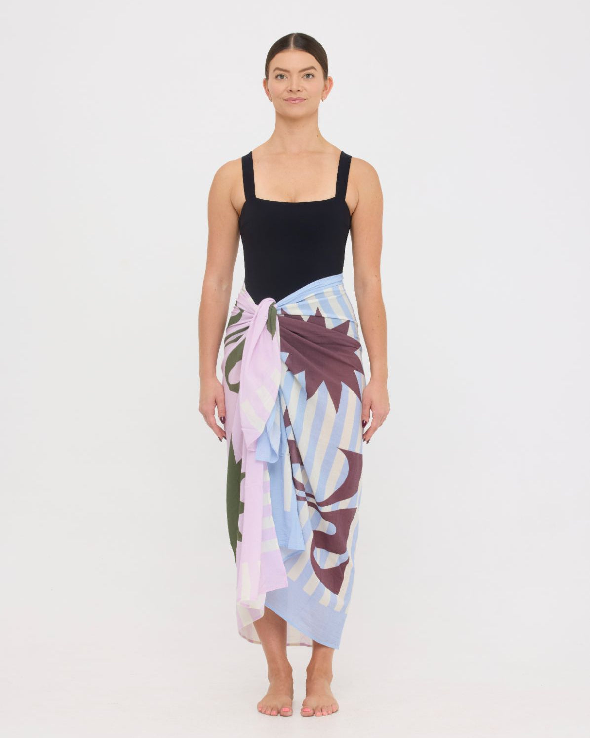 Woman wearing the black HUZZAH Scout sculpting one-piece with a lightweight multi coloured sarong wrapped at the hip, creating a sophisticated swimwear look.
