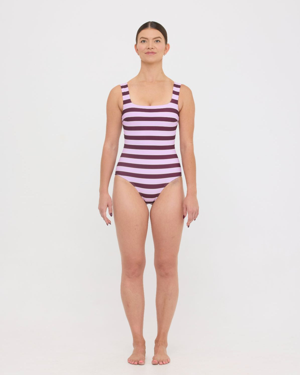 Model wearing the violet and plum mixed stripe HUZZAH Cat one-piece swimsuit with sculpting fit and clean lines, photographed against a plain background