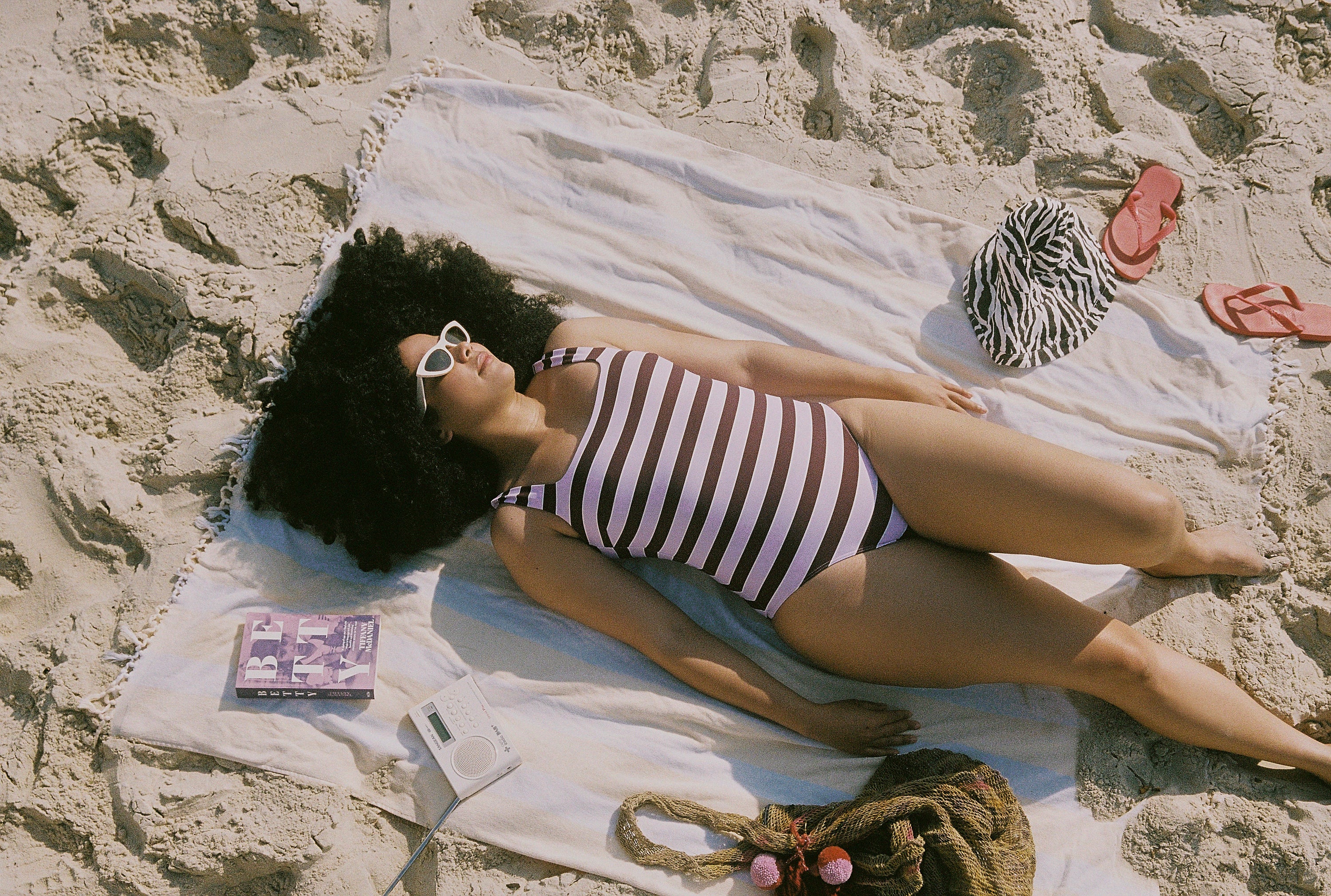 Woman in a HUZZAH mixed striped Cat One Piece swimsuit lying on a towel at the beach with a magazine and bag nearby.