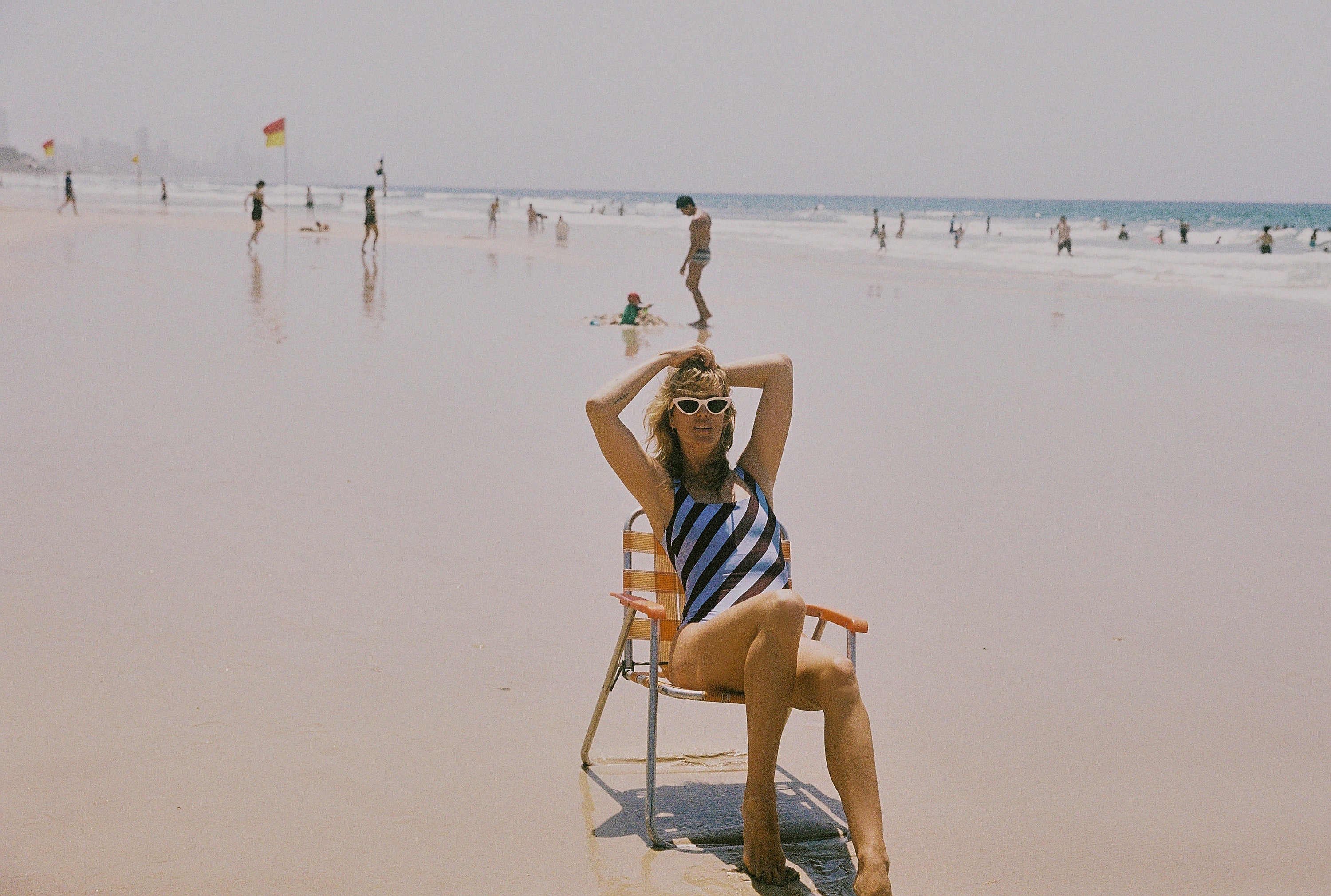 Woman in a HUZZAH dusty blue and plum rising stripe one piece swimsuit sitting on a beach chair at the beach.