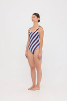 Model wearing the dusty blue and plum rising stripe HUZZAH Cat one-piece swimsuit with sculpting fit and clean lines, photographed against a plain background