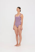 Model wearing the violet and plum mixed stripe HUZZAH Cat one-piece swimsuit with sculpting fit and clean lines, photographed against a plain background