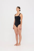 Model wearing the black HUZZAH Scout one-piece swimsuit with sculpting fit and clean lines, photographed against a plain background