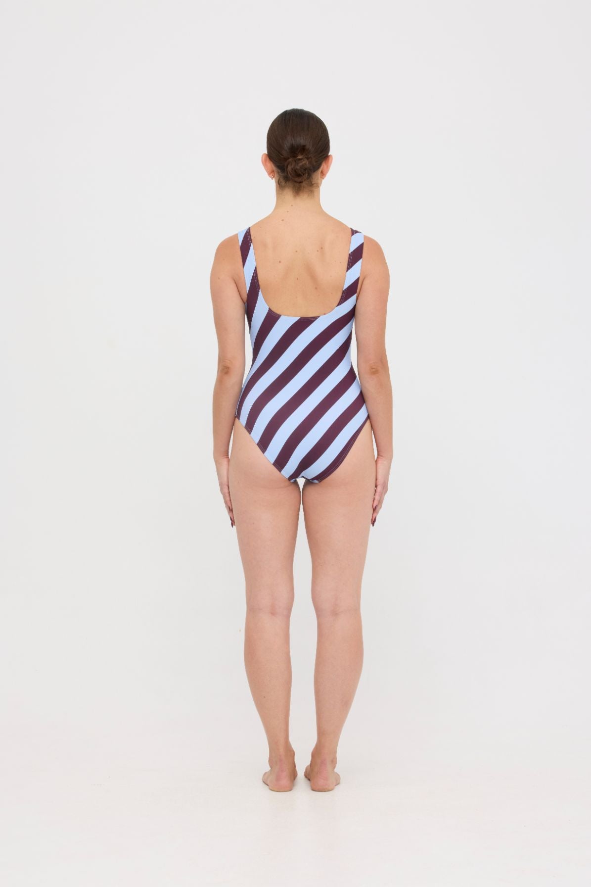 Model wearing the dusty blue and plum rising stripe HUZZAH Cat one-piece swimsuit with sculpting fit and clean lines, photographed against a plain background