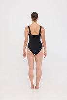 Model wearing the black HUZZAH Scout one-piece swimsuit with sculpting fit and clean lines, photographed against a plain background