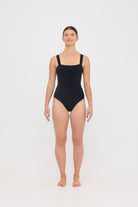 Model wearing the black HUZZAH Scout one-piece swimsuit with sculpting fit and clean lines, photographed against a plain background