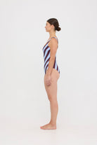 Model wearing the dusty blue and plum rising stripe HUZZAH Cat one-piece swimsuit with sculpting fit and clean lines, photographed against a plain background