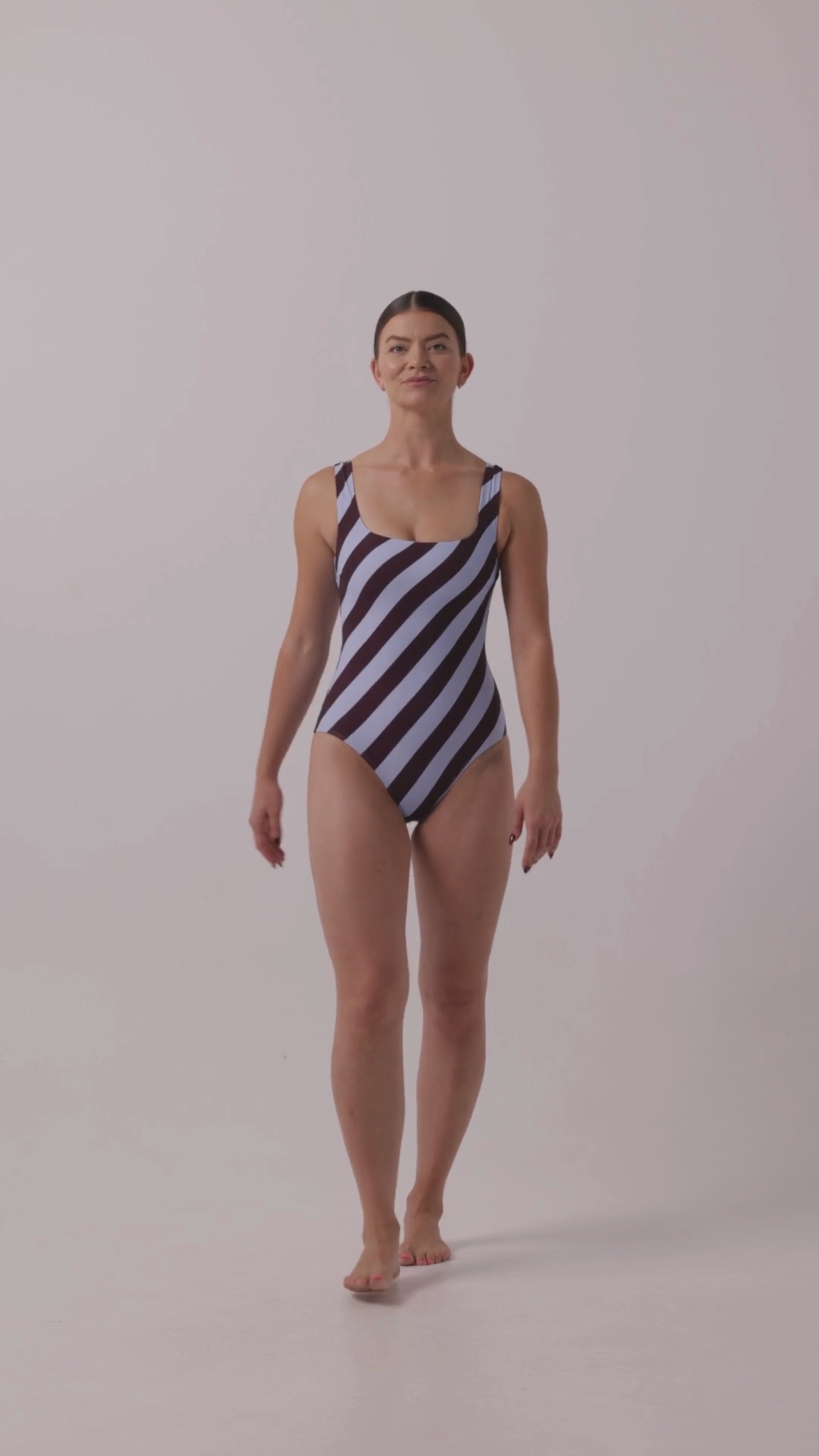 Model wearing the dusty blue and plum rising stripe HUZZAH Cat one-piece swimsuit with sculpting fit and clean lines, photographed against a plain background