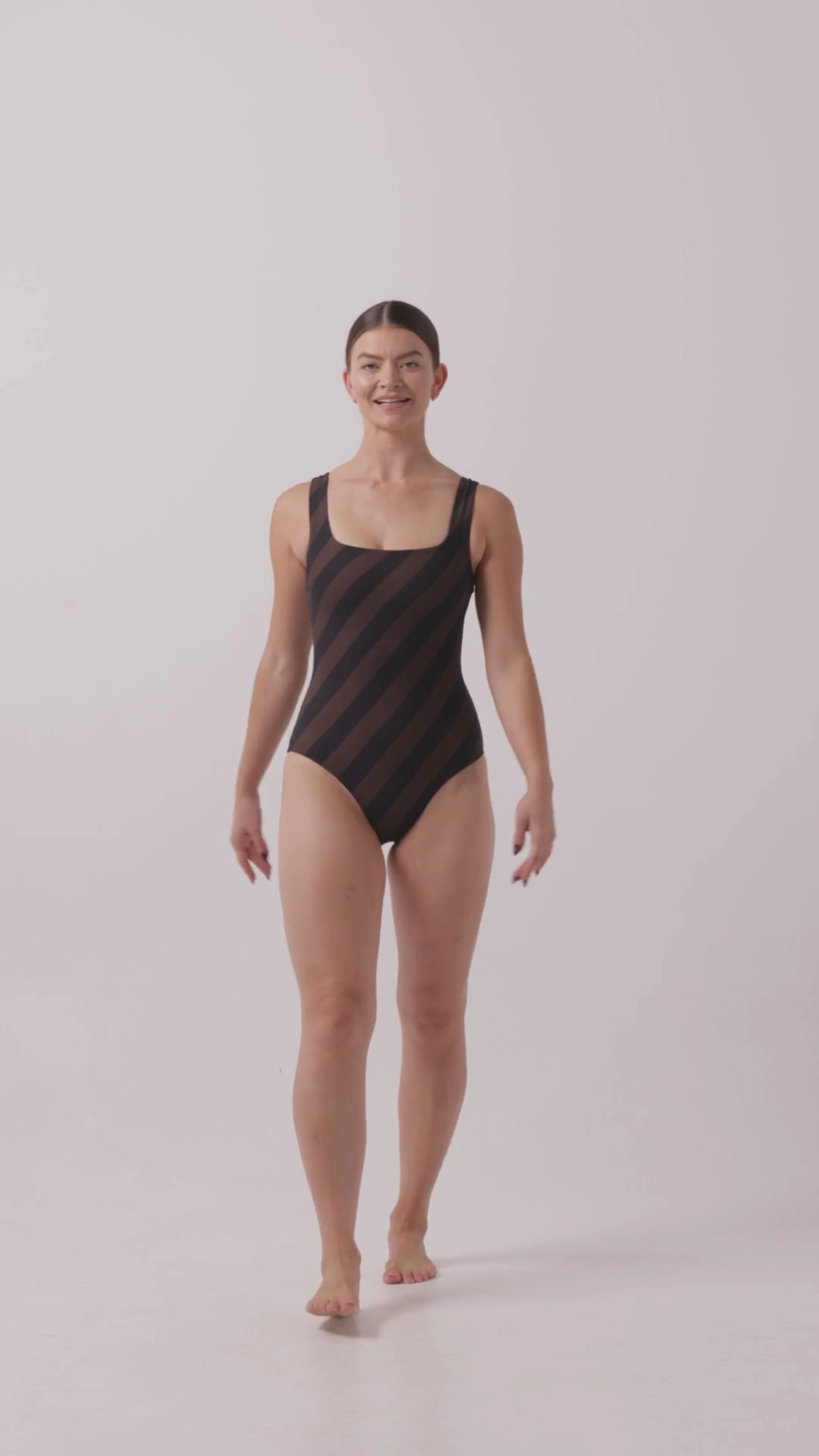 Model wearing the espresso and black rising stripe HUZZAH Cat one-piece swimsuit with sculpting fit and clean lines, photographed against a plain background